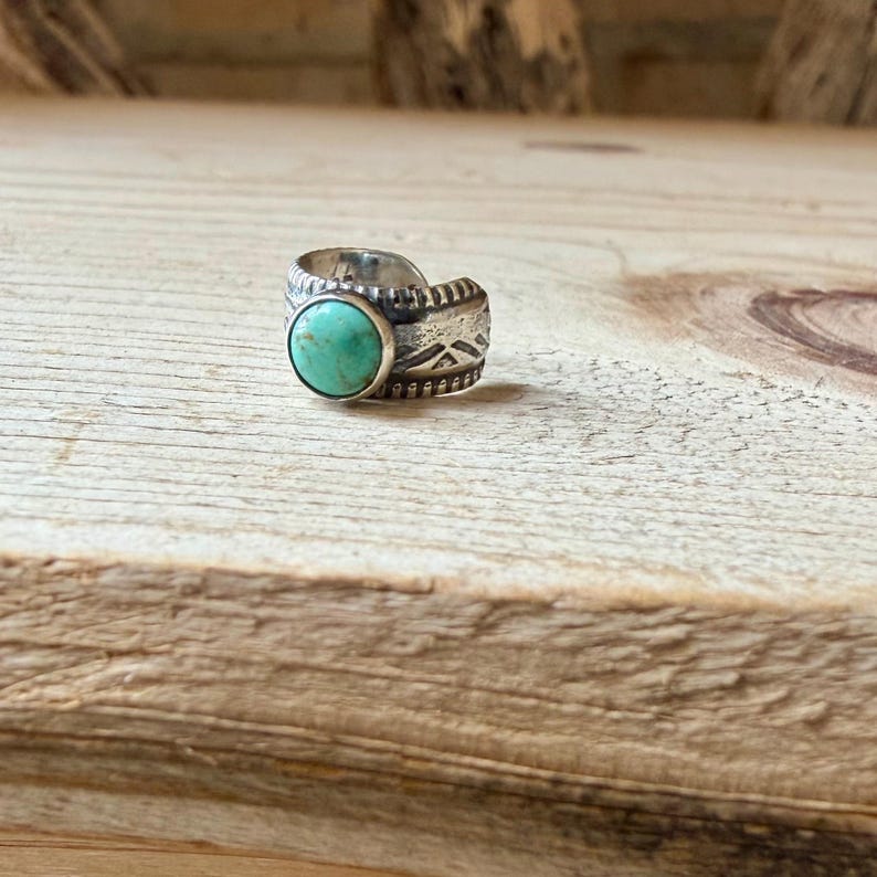 May include: A silver ring featuring a round, polished turquoise stone set in the center. The band is crafted from oxidized silver with a textured surface, displaying etched geometric patterns and a beaded border detail along the edges.