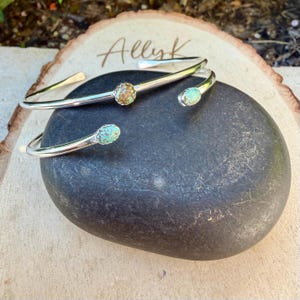 May include: Two silver-toned cuff bracelets with turquoise and brown stone accents. The bracelets are displayed on a dark gray stone, with the name "Allyk" written on a wooden surface in the background. The bracelets have an open design.