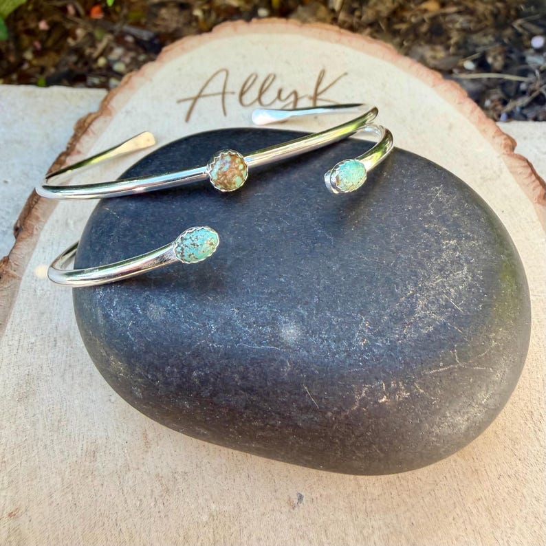 May include: Two silver-toned cuff bracelets with turquoise and brown stone accents. The bracelets are displayed on a dark gray stone, with the name "Allyk" written on a wooden surface in the background. The bracelets have an open design.