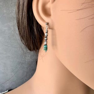 May include: Silver dangle earrings with a post backing. The earrings feature a vertical design with alternating silver beads and small turquoise-colored beads. The earrings are shown on a person's ear.