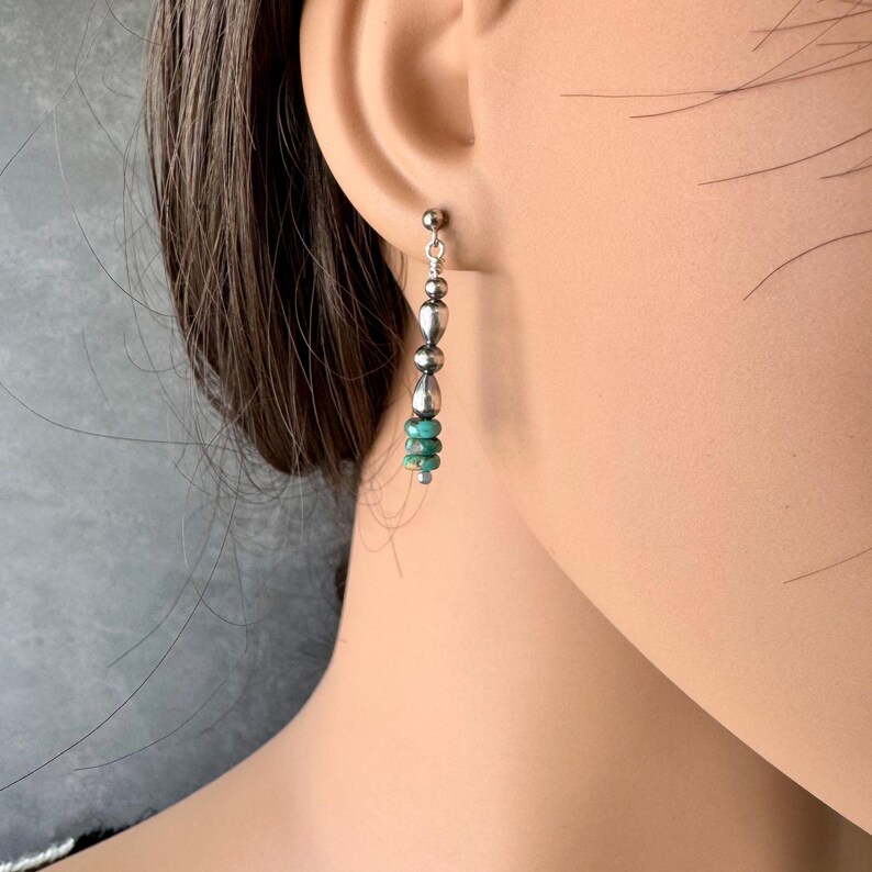 May include: Silver dangle earrings with a post backing. The earrings feature a vertical design with alternating silver beads and small turquoise-colored beads. The earrings are shown on a person's ear.