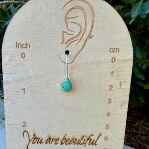 Pu&ograve; includere: Un orecchino con pietra turchese a forma di goccia con montatura in argento, esposto su un cartoncino in legno chiaro con il contorno di un orecchio, segni di misurazione in pollici e centimetri e il testo You are beautiful in basso.