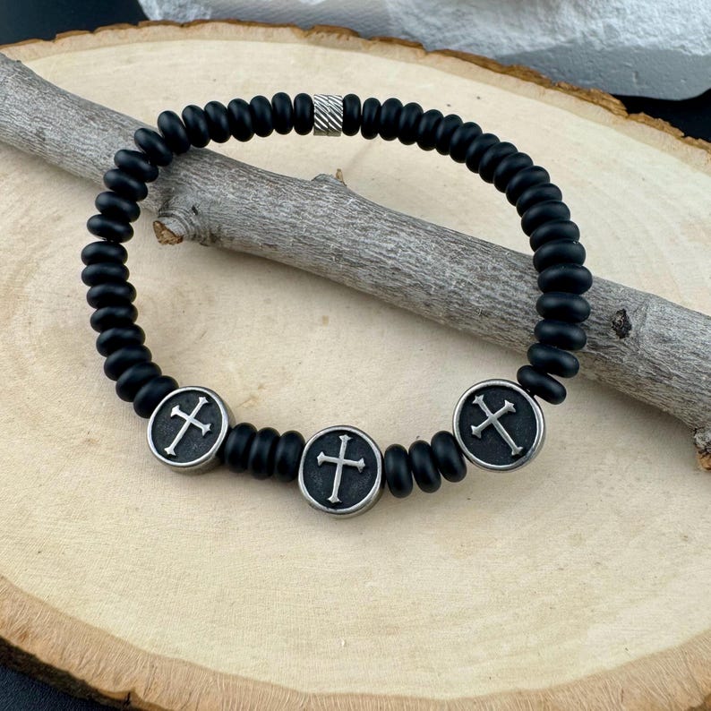 May include: Black beaded bracelet with three silver-toned cross charms. The bracelet features a mix of matte black beads and silver accents. The cross charms are circular with a cross design in the center. The bracelet is displayed on a wooden surface.