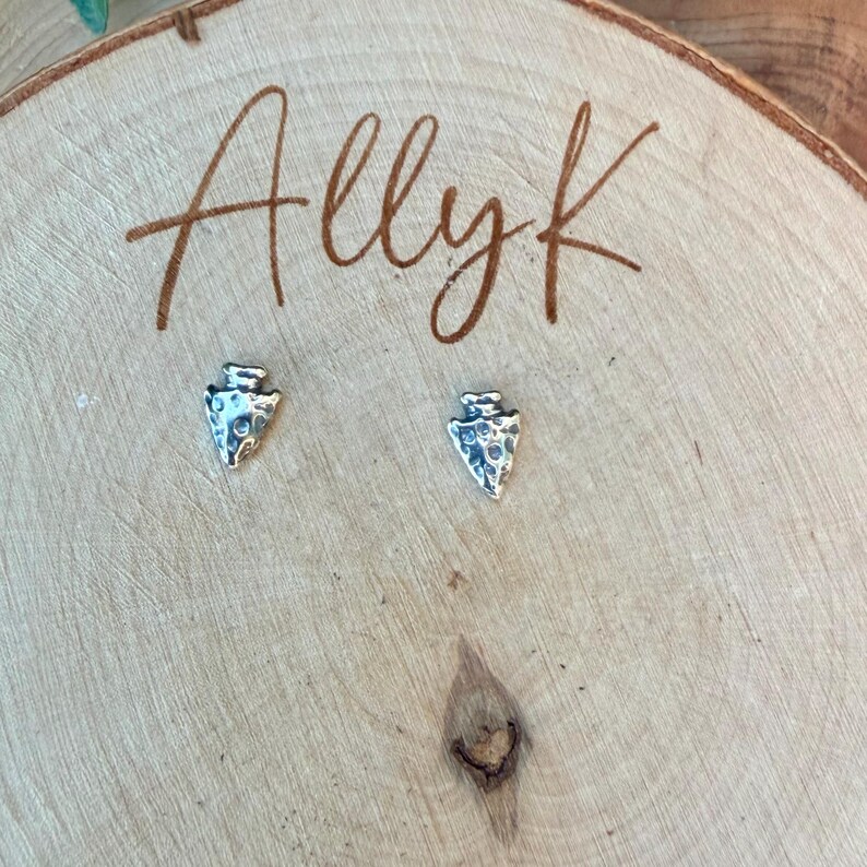 May include: A pair of silver-colored arrowhead stud earrings. The earrings are displayed on a wooden surface with the handwritten text "Allyk" in brown script. The earrings have a textured surface.