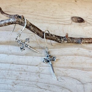 May include: A pair of silver-toned earrings featuring a cross-shaped pendant with ornate, fleur-de-lis style detailing at the top and a pointed, dagger-like base. Each earring hangs from a simple curved silver wire hook for attachment.