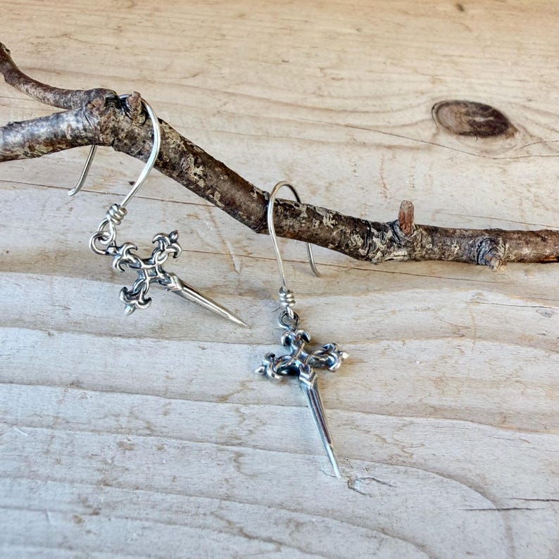 May include: A pair of silver-toned earrings featuring a cross-shaped pendant with ornate, fleur-de-lis style detailing at the top and a pointed, dagger-like base. Each earring hangs from a simple curved silver wire hook for attachment.