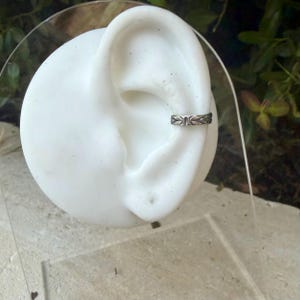 K&ouml;nnte beinhalten: Ein silberfarbener Ohrring in Form einer Manschette mit einem sich wiederholenden, gepr&auml;gten Blattmuster entlang des Bandes. Das Metall weist ein dunkles, oxidiertes Finish in den vertieften Bereichen auf, das die botanischen Details hervorhebt.