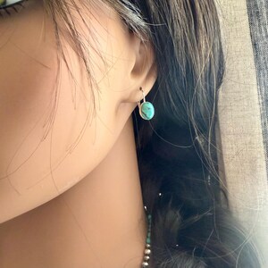 May include: A close-up of a silver earring with a turquoise oval stone. The earring is a simple hoop design, and the turquoise stone has a light blue-green color. The earring is worn on the ear.