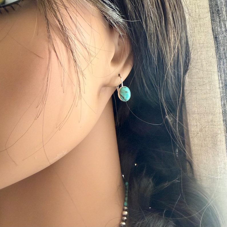 May include: A close-up of a silver earring with a turquoise oval stone. The earring is a simple hoop design, and the turquoise stone has a light blue-green color. The earring is worn on the ear.