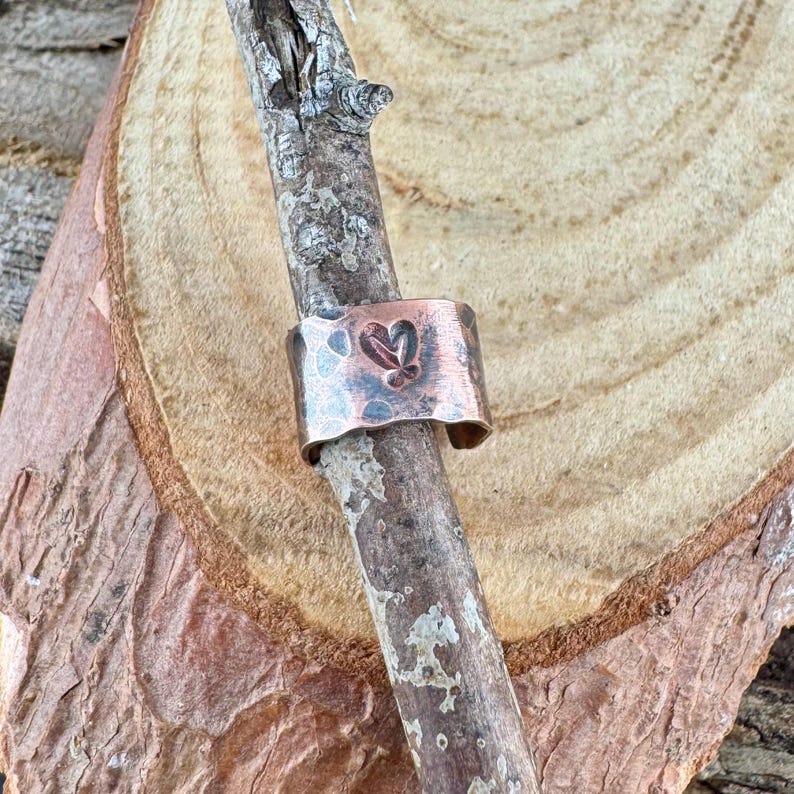 May include: A copper-colored ring with a hammered texture and a heart-shaped cutout. The ring is displayed on a small branch against a natural wood background. The ring's band is approximately 0.75 inches wide.