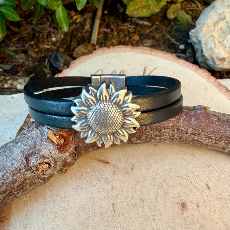 May include: Black leather wrap bracelet with a silver sunflower charm. The bracelet features two bands of black leather and a silver sunflower charm with detailed petals. The clasp is silver and the bracelet is displayed on a wooden surface.