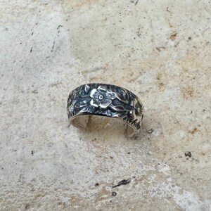 May include: A silver ring with a floral design. The ring features a band covered in detailed, raised flower and leaf patterns. The metal has a slightly oxidized appearance, enhancing the intricate details of the design. The ring is set against a neutral background.