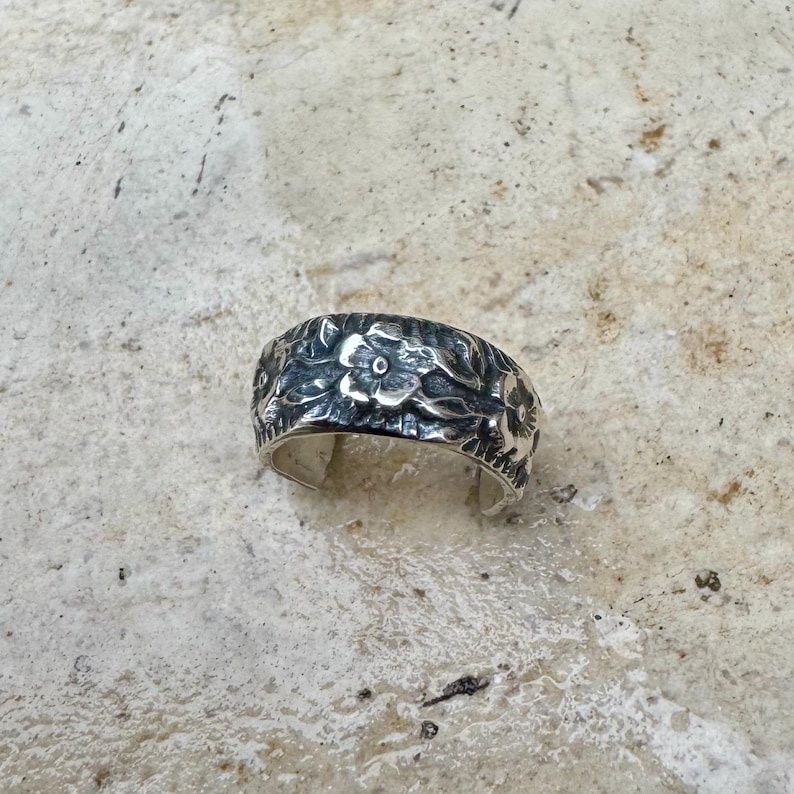 May include: A silver ring with a floral design. The ring features a band covered in detailed, raised flower and leaf patterns. The metal has a slightly oxidized appearance, enhancing the intricate details of the design. The ring is set against a neutral background.