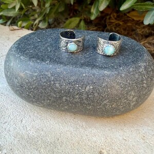 May include: Two open-ended silver rings featuring intricate etched patterns and a single round, light blue cabochon stone set in the center of each band. The rings have a polished finish and a wide, adjustable design with a visible gap at the back.