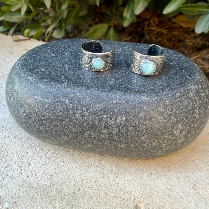 May include: Two open-ended silver rings featuring intricate etched patterns and a single round, light blue cabochon stone set in the center of each band. The rings have a polished finish and a wide, adjustable design with a visible gap at the back.