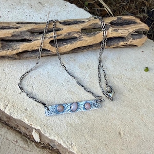 May include: A silver-toned metal necklace featuring a rectangular bar pendant with an etched scrollwork pattern. The pendant is set with three smooth, oval-shaped pink stones arranged horizontally. The chain consists of rectangular links.