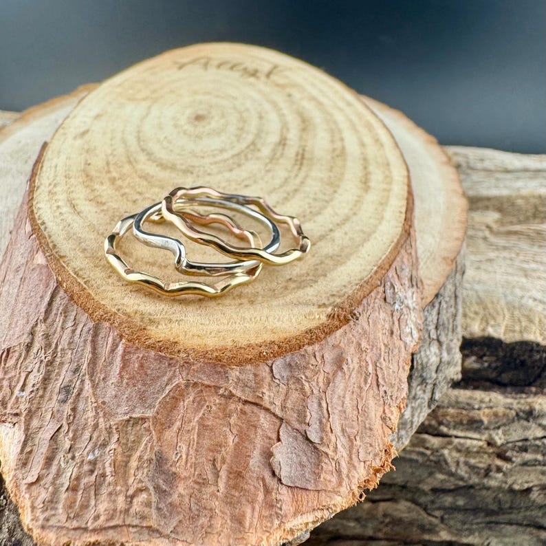 Puede incluir: Tres anillos con bordes ondulados en tonos oro, plata y oro rosa se exhiben sobre una rodaja de madera. Los anillos est&aacute;n apilados, mostrando su dise&ntilde;o &uacute;nico y org&aacute;nico y su brillo met&aacute;lico. La rodaja de madera tiene un borde de corteza natural.