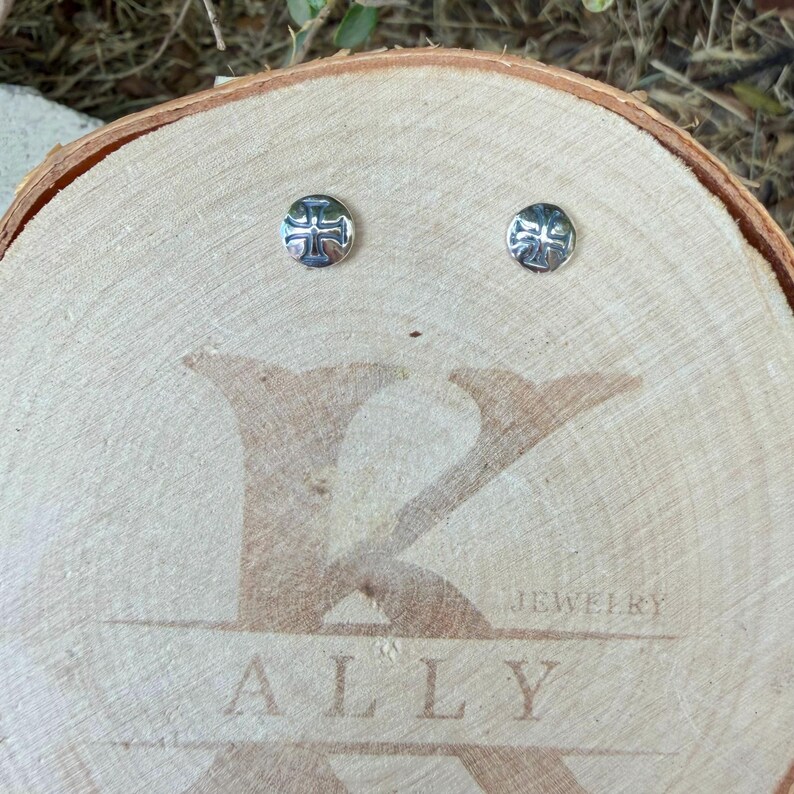 May include: A pair of small, round, silver-toned stud earrings, each featuring an embossed cross design. The earrings are displayed on a circular wooden slice engraved with the text K ALLY JEWELRY in a large, stylized font.
