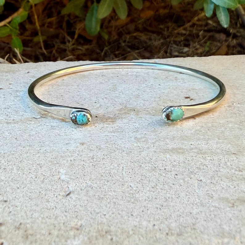 May include: A silver cuff bracelet featuring a smooth, polished metal band that is open at the front. Each end of the bracelet is capped with a small, oval-shaped turquoise stone, displaying natural blue and green colour variations.