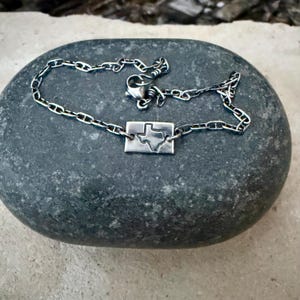 May include: A silver-toned necklace with a rectangular pendant featuring a cutout of the state of Texas. The necklace chain has a link design. The jewelry is displayed on a gray stone.