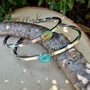 May include: Two silver cuff bracelets featuring twisted metal bands and oval-shaped cabochon stones. One bracelet displays a mottled green and brown stone, while the other features a bright turquoise stone, both set in serrated metal bezels.