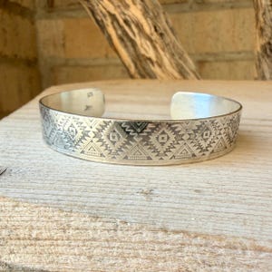May include: A silver cuff bracelet featuring a repeating geometric pattern of etched triangles, diamonds, and horizontal lines. The metal has a polished finish with darkened recessed areas that highlight the intricate stamped design across the band.