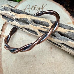 May include: A copper-toned twisted metal bracelet. The bracelet is made of two strands of metal that are intertwined. The bracelet is resting on a piece of wood. The word "Ally K" is written on the wood.