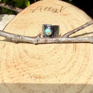 May include: A wide, silver-toned ring with a turquoise stone set in the center. The ring is displayed on a wooden slice with the name "Allyk" etched into the wood. A small branch rests in front of the ring.