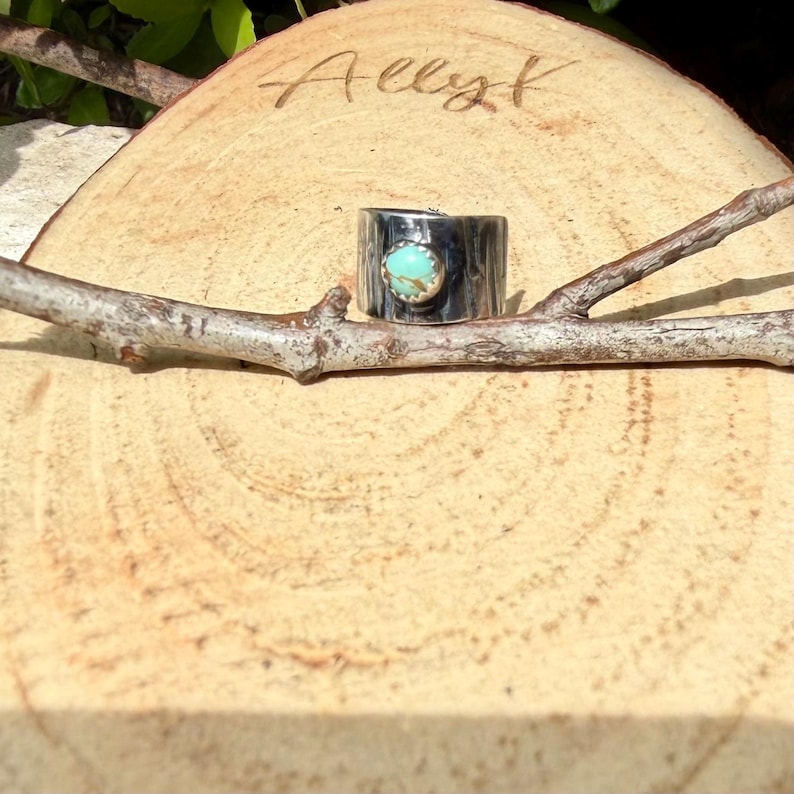 May include: A wide, silver-toned ring with a turquoise stone set in the center. The ring is displayed on a wooden slice with the name "Allyk" etched into the wood. A small branch rests in front of the ring.