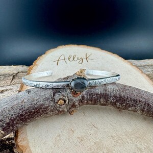 May include: A silver cuff bracelet with a textured band and an oval, dark gray gemstone set in a gold-colored bezel. The bracelet rests on a wooden branch, with the name "AllyK" written on a wooden slice in the background.