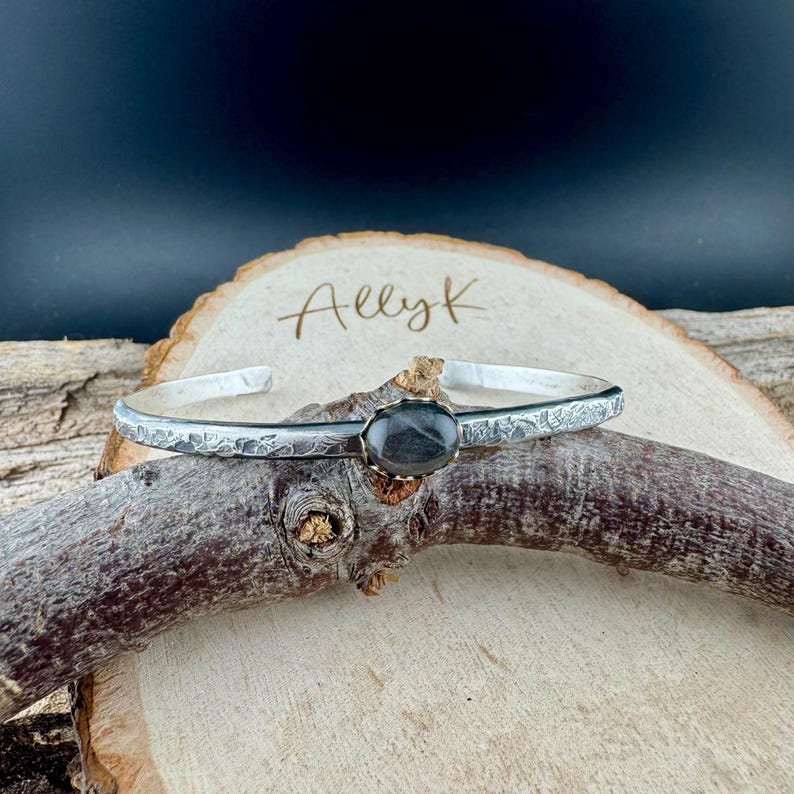 May include: A silver cuff bracelet with a textured band and an oval, dark gray gemstone set in a gold-colored bezel. The bracelet rests on a wooden branch, with the name "AllyK" written on a wooden slice in the background.