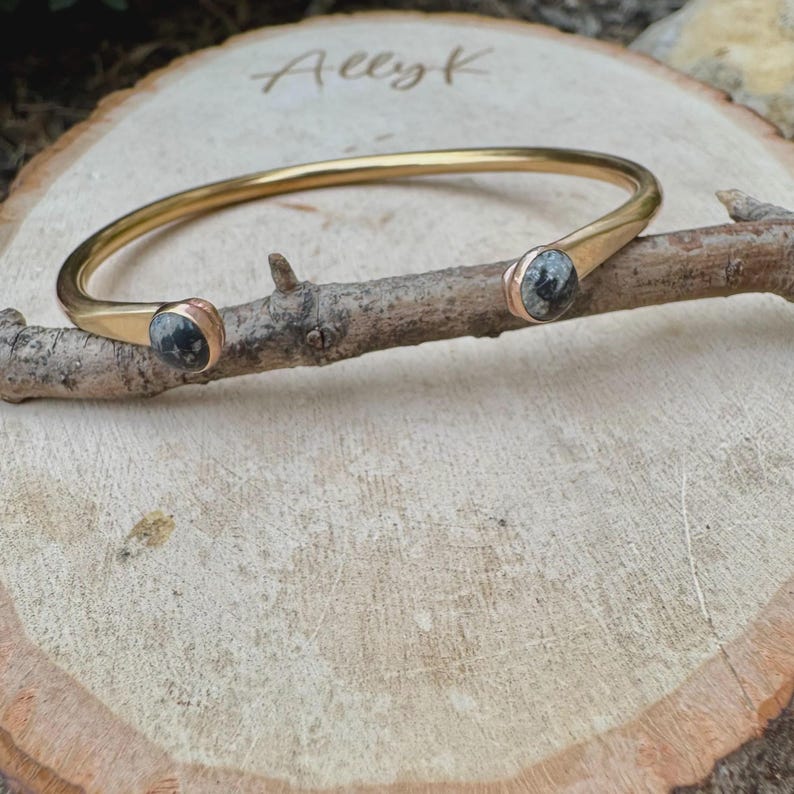 May include: A gold-colored metal bracelet with a circular design. The bracelet features two gray and black stone accents at the ends. The bracelet is resting on a wooden branch, which is placed on a wooden surface.