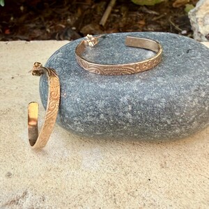 May include: Two gold-colored hoop earrings with an engraved floral pattern. The earrings are displayed on a gray stone. The earrings have a polished finish and a classic design, suitable for everyday wear.
