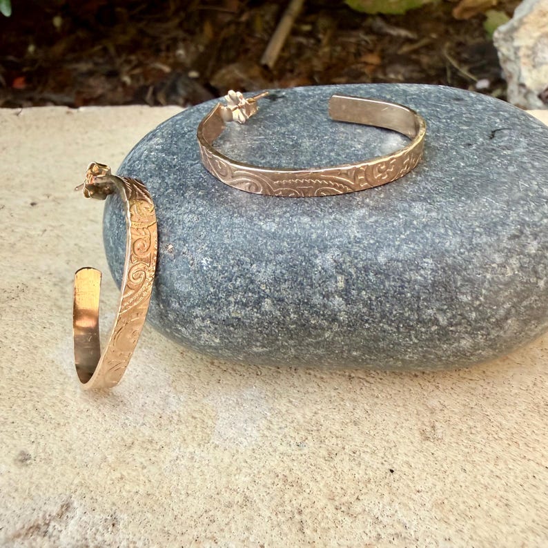 May include: Two gold-colored hoop earrings with an engraved floral pattern. The earrings are displayed on a gray stone. The earrings have a polished finish and a classic design, suitable for everyday wear.