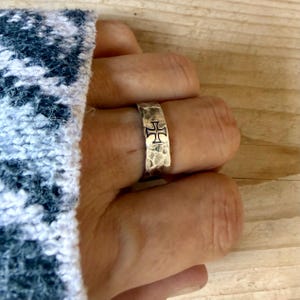 May include: A silver-toned ring with a hammered, textured surface and a prominent black cross symbol engraved on the front. The band has a wide, flat profile and a metallic finish, showcasing a distinct contrast between the dark cross and the silver metal.