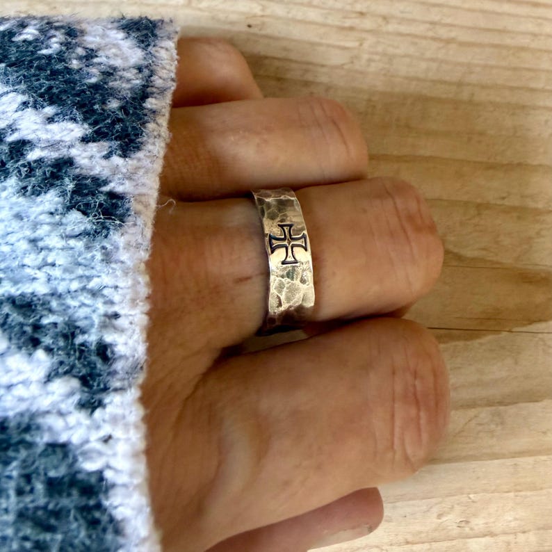 May include: A silver-toned ring with a hammered, textured surface and a prominent black cross symbol engraved on the front. The band has a wide, flat profile and a metallic finish, showcasing a distinct contrast between the dark cross and the silver metal.