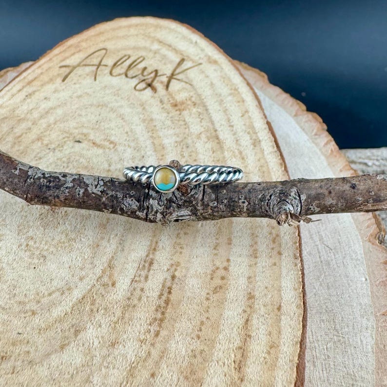 May include: A silver ring with a braided band and a central turquoise and yellow stone. The ring is displayed on a small branch against a wooden background.