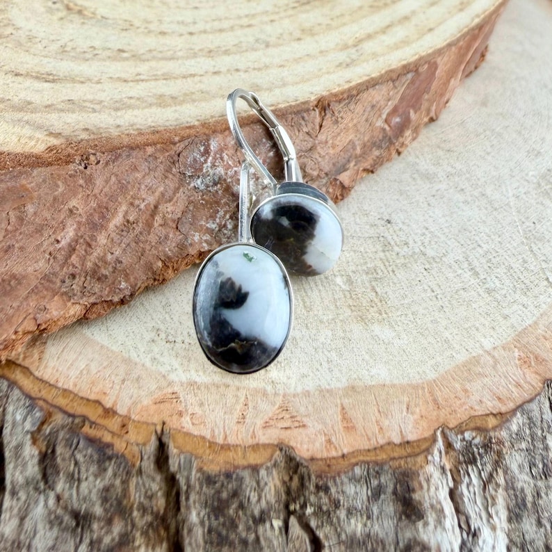 May include: A pair of silver drop earrings with oval-shaped, black and white patterned stones. The earrings are set against a natural wood background, showcasing the contrast between the polished metal and the organic texture.