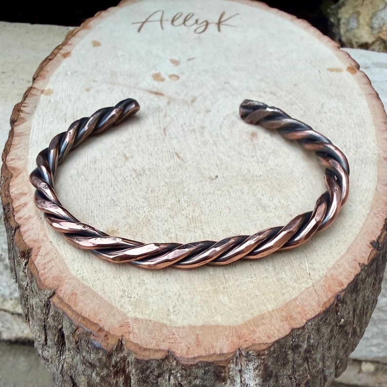 May include: A copper-colored, twisted metal bracelet. The bracelet is open-ended and sits on a wooden surface. The word "Allyk" is written in cursive on the wood. The bracelet has a textured, braided appearance.