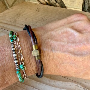 May include: A layered bracelet set consisting of a strand of small turquoise beads and a silver-toned chain, paired with a dark brown leather cord bracelet featuring a central hexagonal brass-toned metal clasp component.