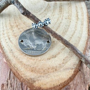 Puede incluir: Un anillo plateado con un dise&ntilde;o de cuentas descansa sobre una peque&ntilde;a rama, encima de una moneda plateada con una imagen de b&uacute;falo. La moneda est&aacute; marcada con "FIVE CENTS" y "UNITED STATES OF AMERICA". El anillo y la moneda se exhiben sobre una superficie de madera.