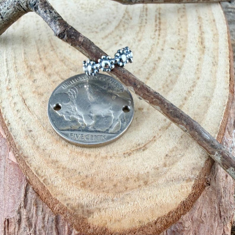 Puede incluir: Un anillo plateado con un dise&ntilde;o de cuentas descansa sobre una peque&ntilde;a rama, encima de una moneda plateada con una imagen de b&uacute;falo. La moneda est&aacute; marcada con "FIVE CENTS" y "UNITED STATES OF AMERICA". El anillo y la moneda se exhiben sobre una superficie de madera.