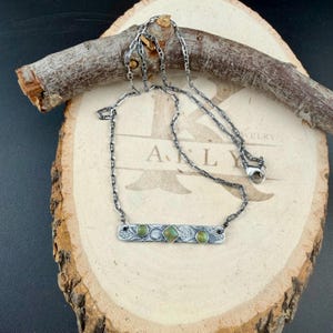 May include: A silver-toned necklace featuring a rectangular bar pendant with an intricate etched pattern. The bar is set with two small round green stones on the sides and a central square green stone. The chain is a dark, oxidized silver link style.