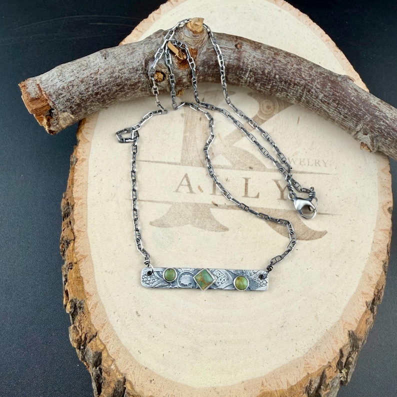 May include: A silver-toned necklace featuring a rectangular bar pendant with an intricate etched pattern. The bar is set with two small round green stones on the sides and a central square green stone. The chain is a dark, oxidized silver link style.