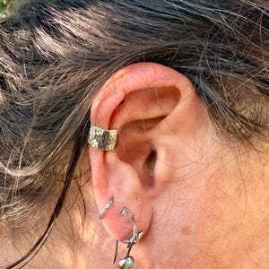 Puede incluir: Un pendiente tipo ear cuff de plata con superficie texturizada y un patr&oacute;n de cruz oscura estampado. El pendiente se sit&uacute;a en el cart&iacute;lago superior de la oreja, acompa&ntilde;ado de un peque&ntilde;o aro de plata y un pendiente colgante con piedra verde.