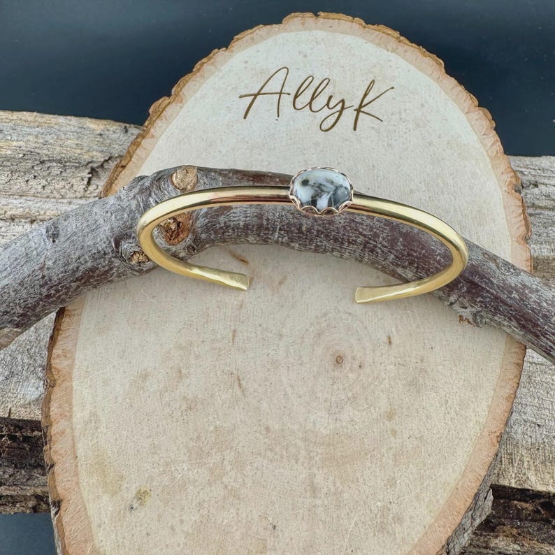 May include: A gold-colored cuff bracelet with a central, round stone featuring a black and white pattern. The bracelet is displayed on a wooden slice with the name "Allyk" written on it.