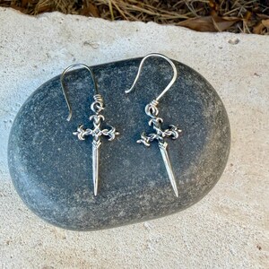 May include: A pair of silver-toned drop earrings featuring a detailed cross design with a pointed, dagger-like base. Each earring hangs from a curved wire hook, showcasing intricate metalwork with darkened recessed areas to highlight the ornate shape.