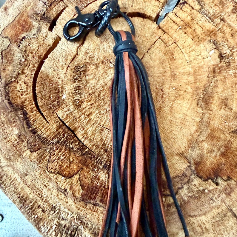 May include: A black leather keychain with a black clasp and a tassel of black and brown leather strips. The keychain is attached to a metal ring.