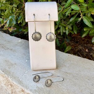 May include: Two pairs of silver-toned earrings featuring a circular, sunburst-patterned design with radiating lines. Each earring is attached to a long, curved hook wire. The metal has a matte, slightly oxidized finish with detailed radial grooves.
