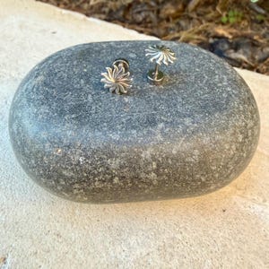 May include: Two silver-toned stud earrings featuring a sunburst design with radiating rays. The earrings have a metallic finish and a post-back mechanism, displayed on a smooth, dark gray, oval-shaped stone surface.